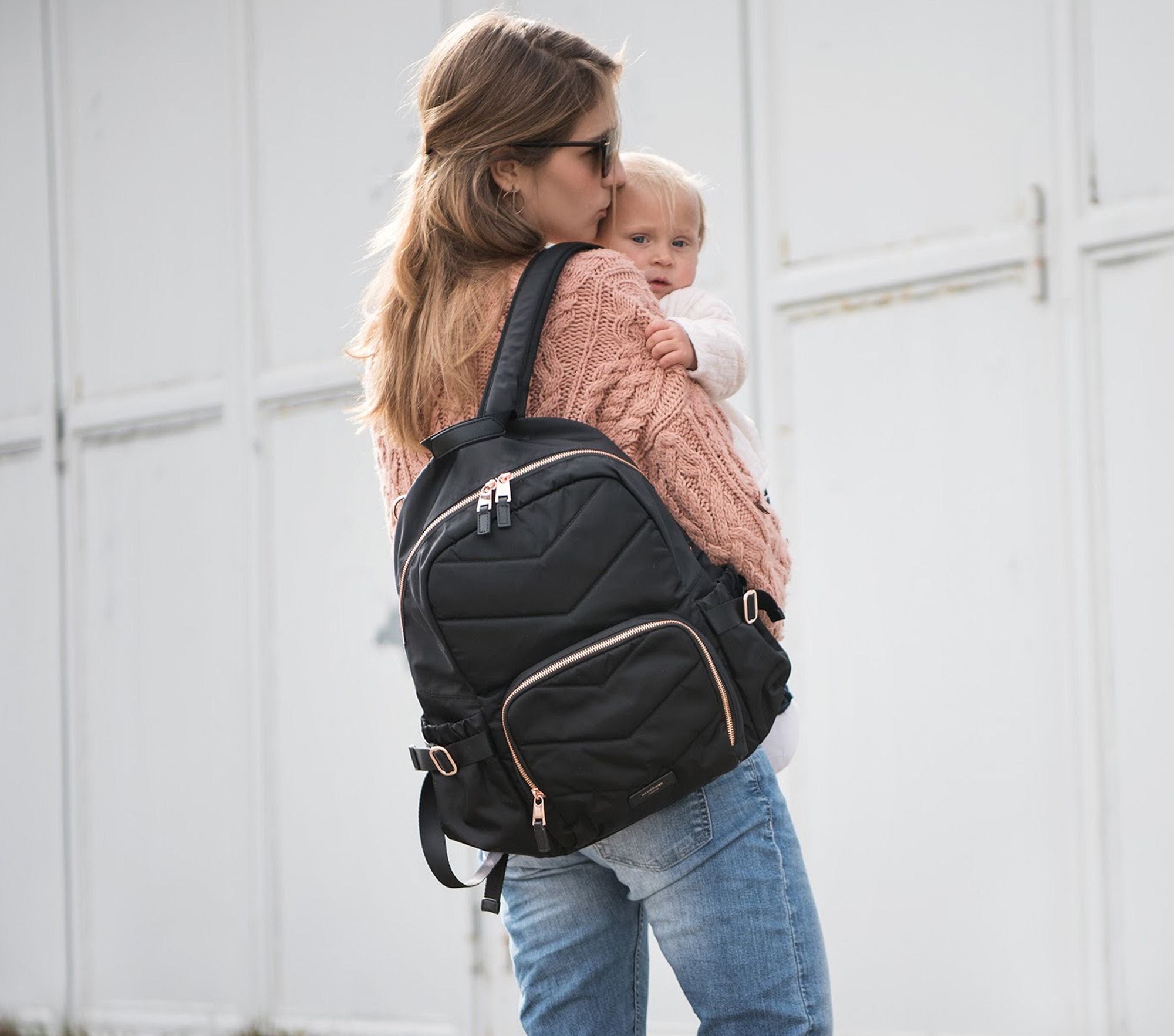 A MOM IS HOLDING HER BABY WHILE CARRYING A DIAPER BAG BACKPACK