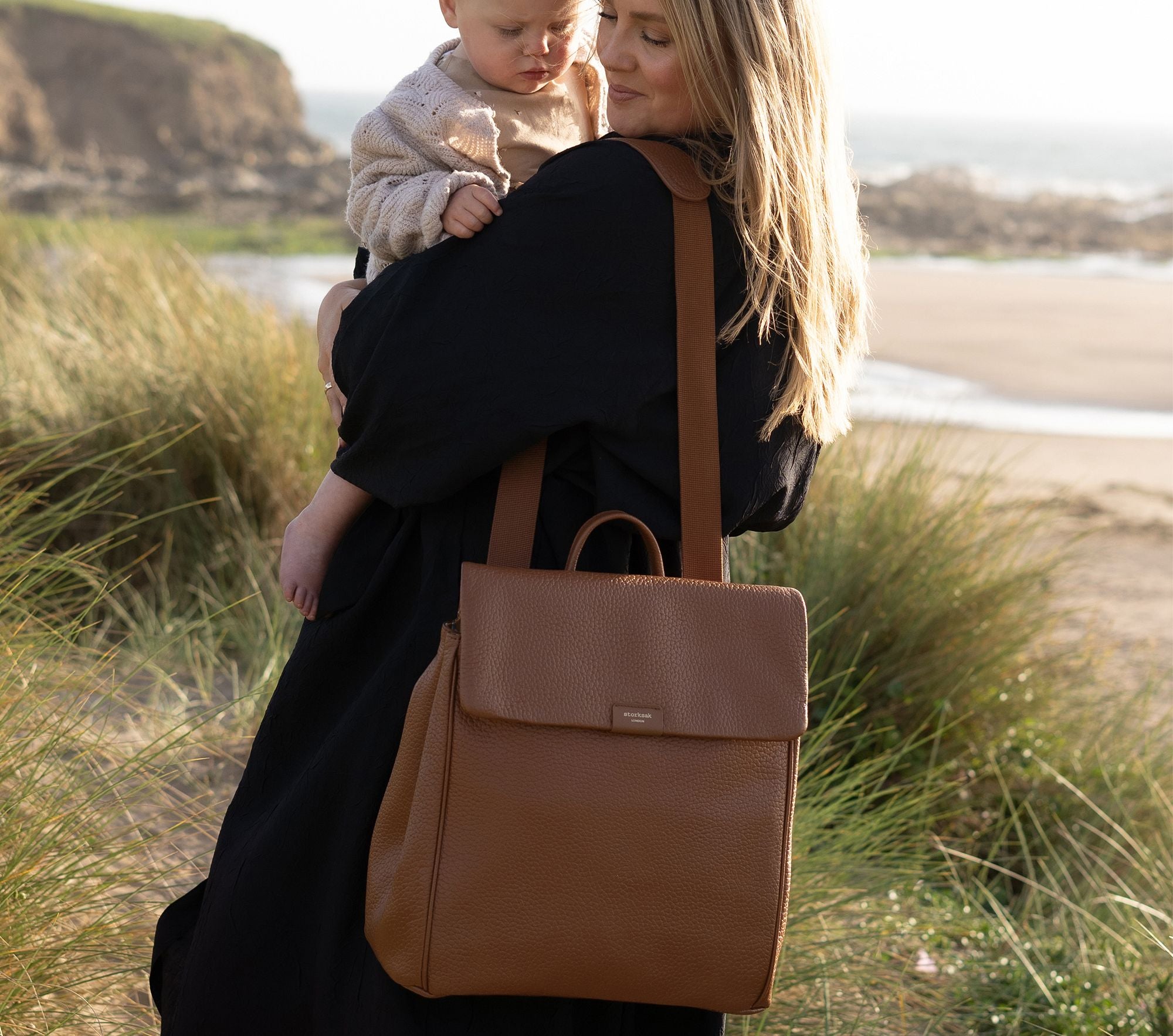 A MOM IS HOLDING HER BABY WHILE CARRYING A LEATHER DIAPER BAG BACKPACK