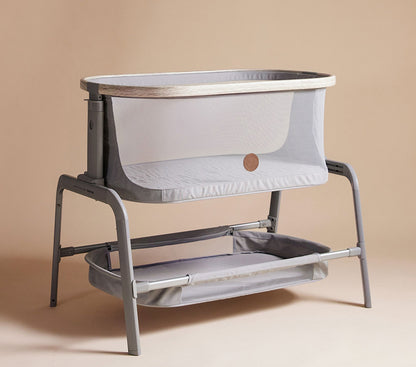angle view of A sleek and modern Maxi-Cosi® Iora newborn Bassinet in a soft gray color, featuring a mesh fabric for breathability and a sturdy metal frame. The bassinet has a lower storage shelf, ideal for keeping baby essentials within reach.