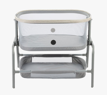 A side view of the Maxi-Cosi® Iora newborn Bassinet, showcasing its sleek design with breathable mesh sides and soft gray fabric. The bassinet includes a convenient lower storage shelf for easy access to baby essentials.
