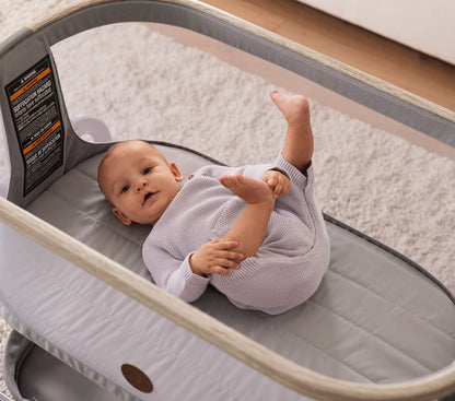 A baby lying comfortably in a Maxi-Cosi® Iora newborn Bassinet, dressed in a light purple onesie. The bassinet's breathable mesh sides and soft gray padding provide a safe and cozy environment for the baby.